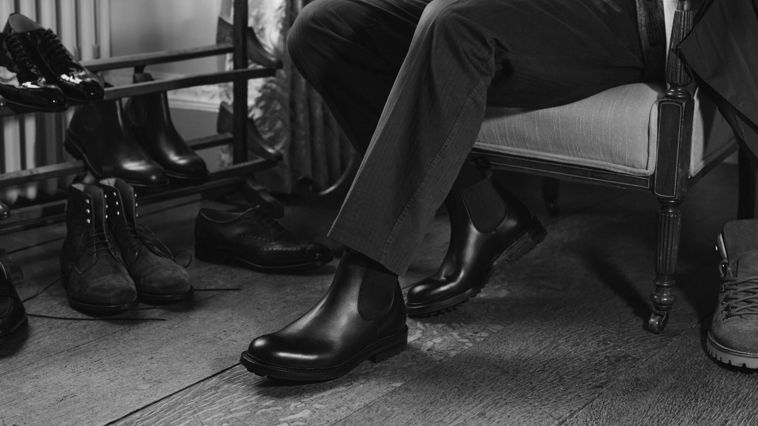 Close-up of a person sitting in an armchair wearing black leather boots, surrounded by shelves and pairs of leather shoes.