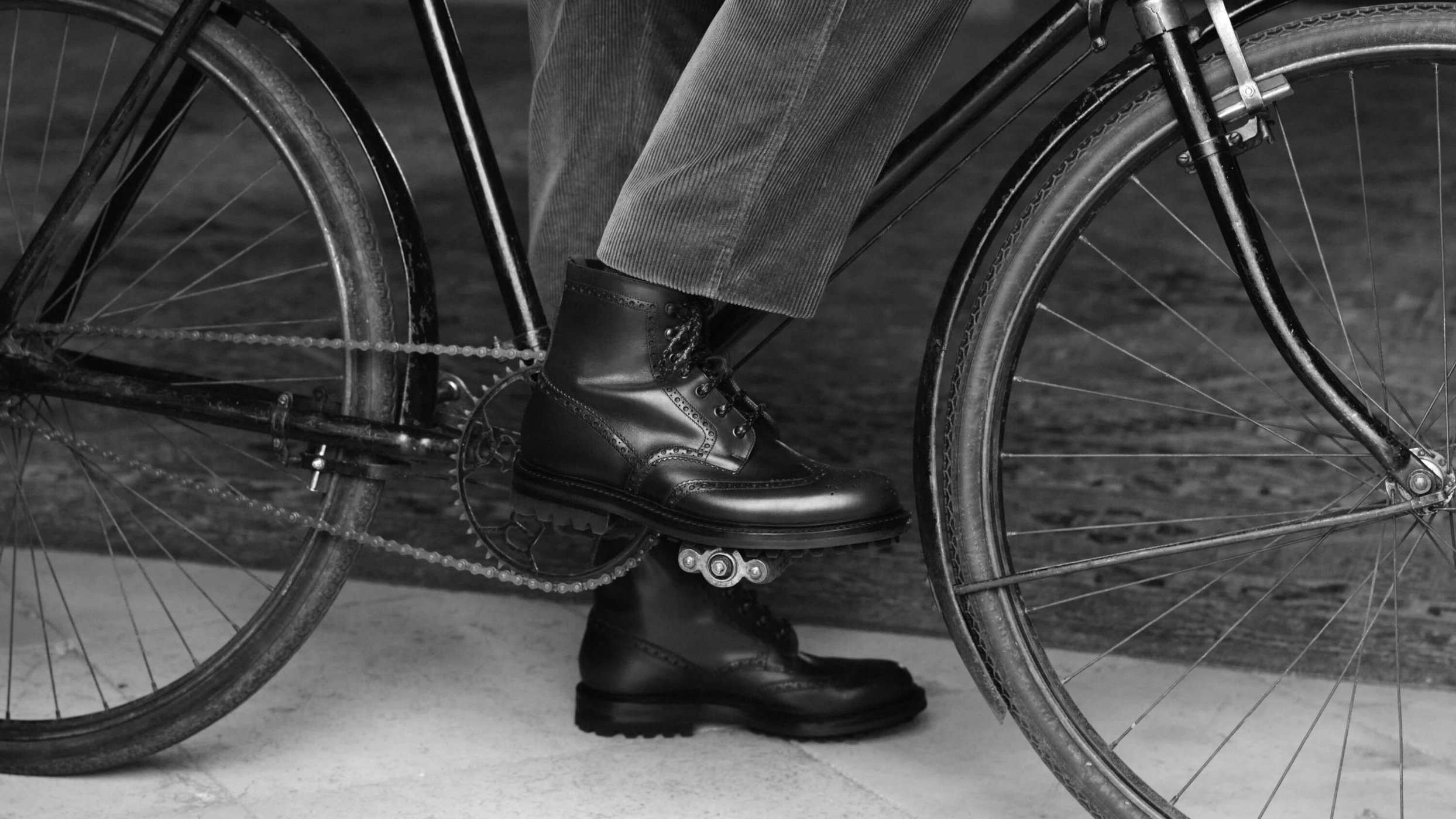 Close-up of black leather boots worn by a person riding a vintage bicycle in a black and white photograph.