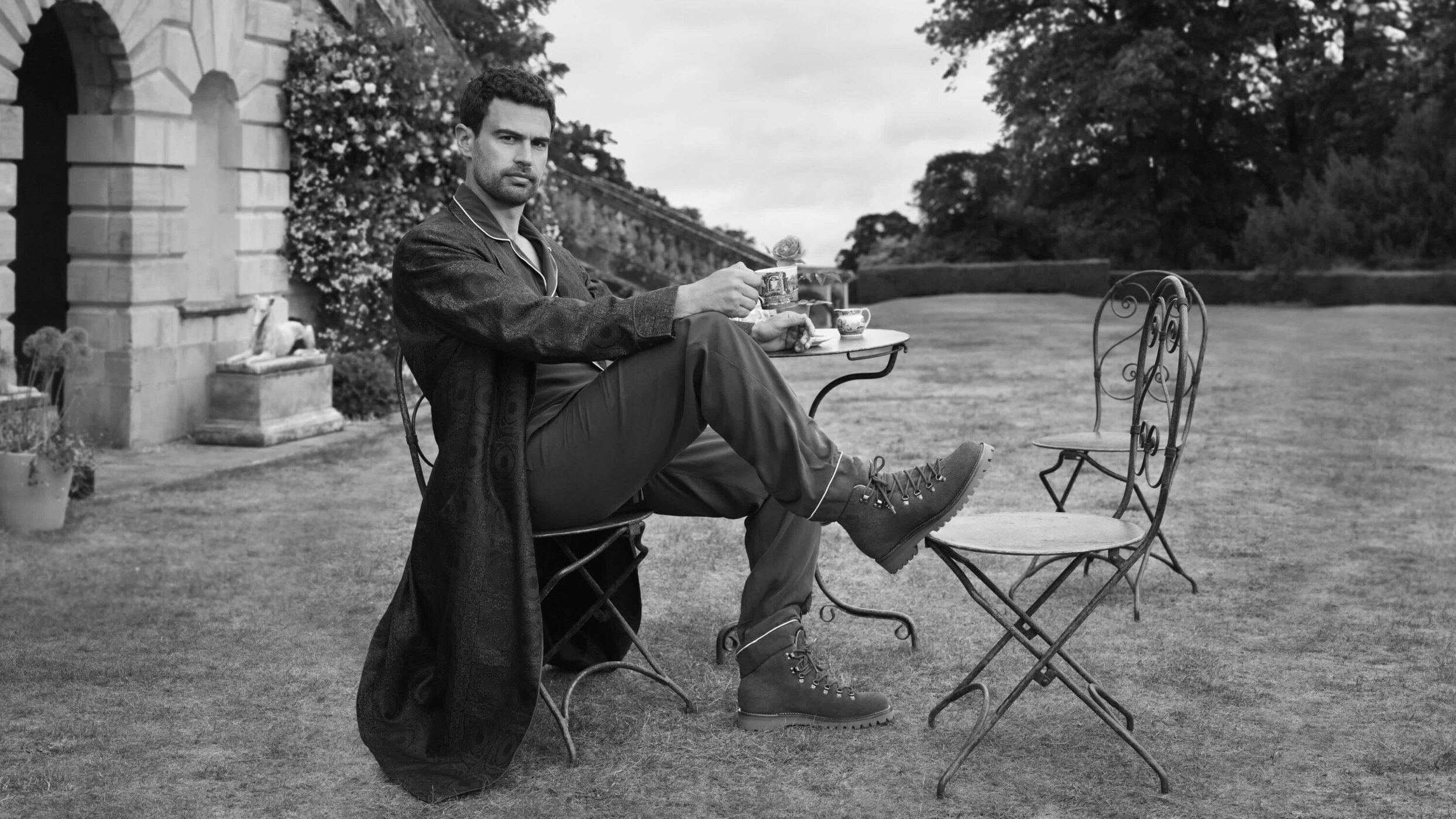 Theo James seated outdoors at a small round table wearing a patterned robe and boots in a black and white campaign photograph.