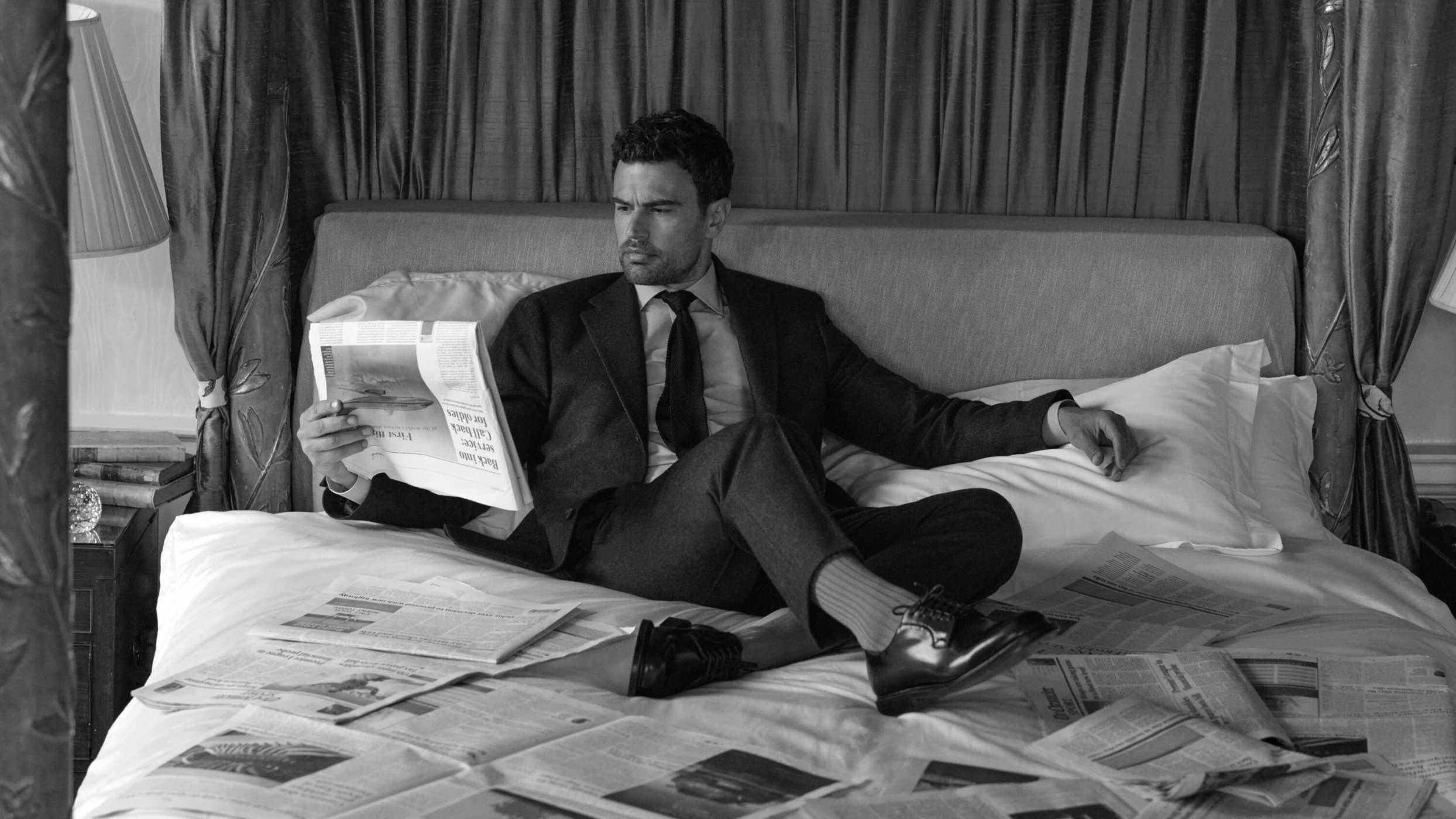 Man in a suit seated on a bed surrounded by newspapers holding one up to read, shot in black and white.