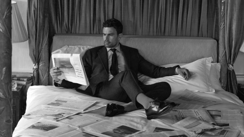 Man in a suit seated on a bed surrounded by newspapers holding one up to read, shot in black and white.