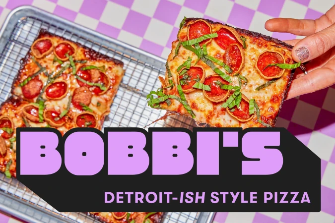 Hand lifting a square pepperoni pizza slice topped with basil from a wire cooling rack against a purple checkerboard background.