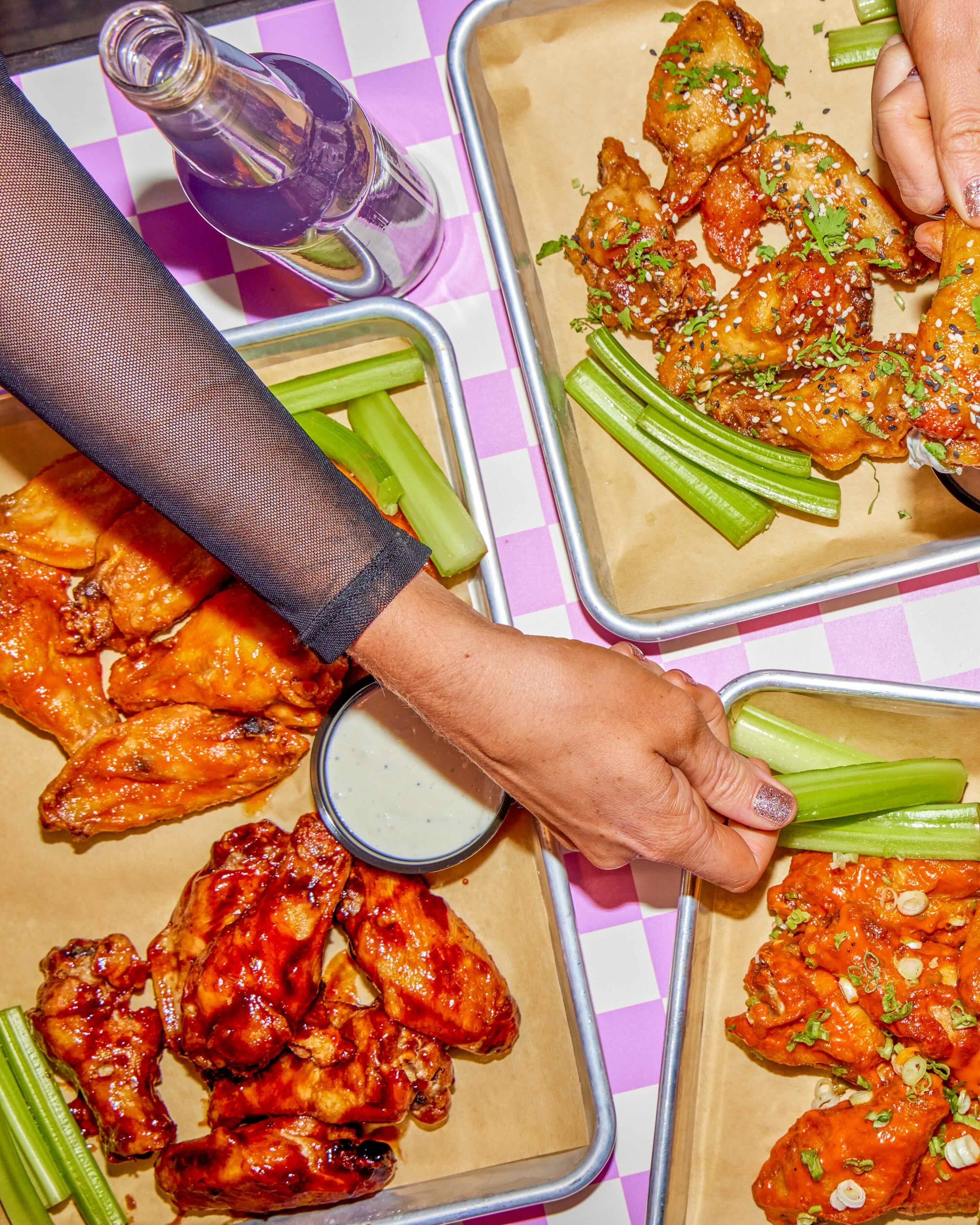 Overhead shot of multiple metal trays filled with glazed chicken wings, celery sticks, and a glass bottle on a pink checked surface.