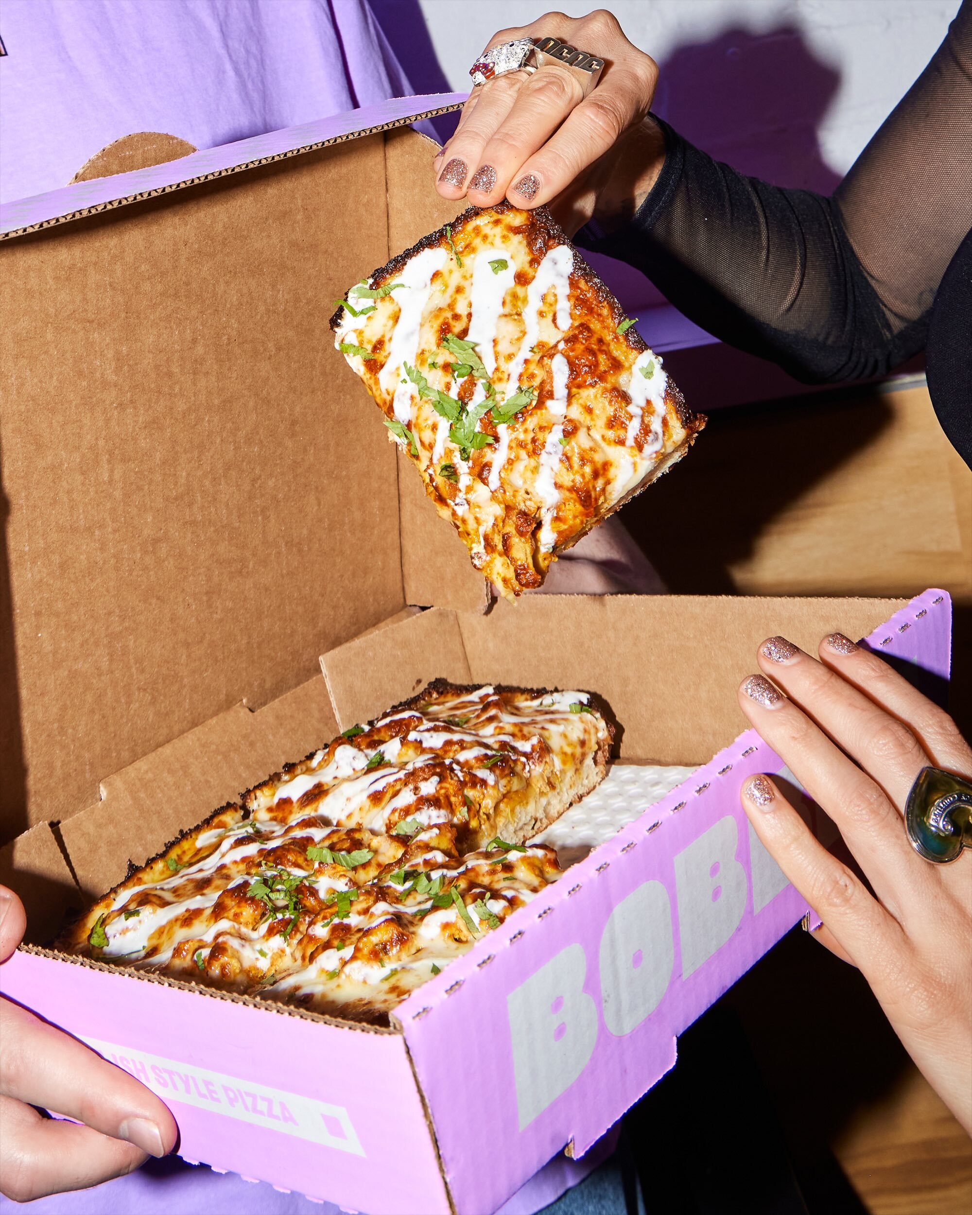 Close-up of hands lifting a slice of cheesy pizza from an open purple branded pizza box.