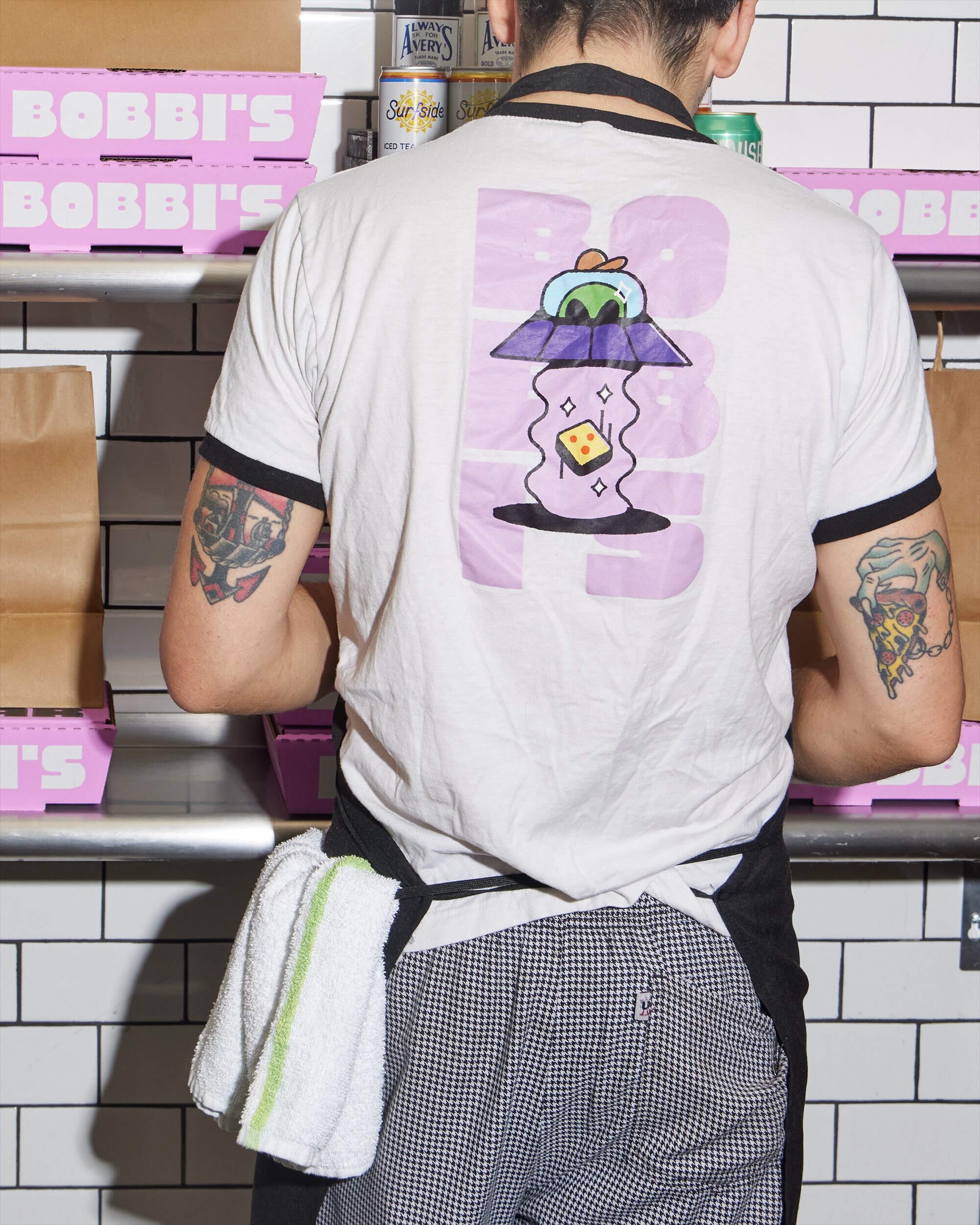 Person wearing ringer t-shirt with UFO graphic and houndstooth trousers in a commercial kitchen setting.