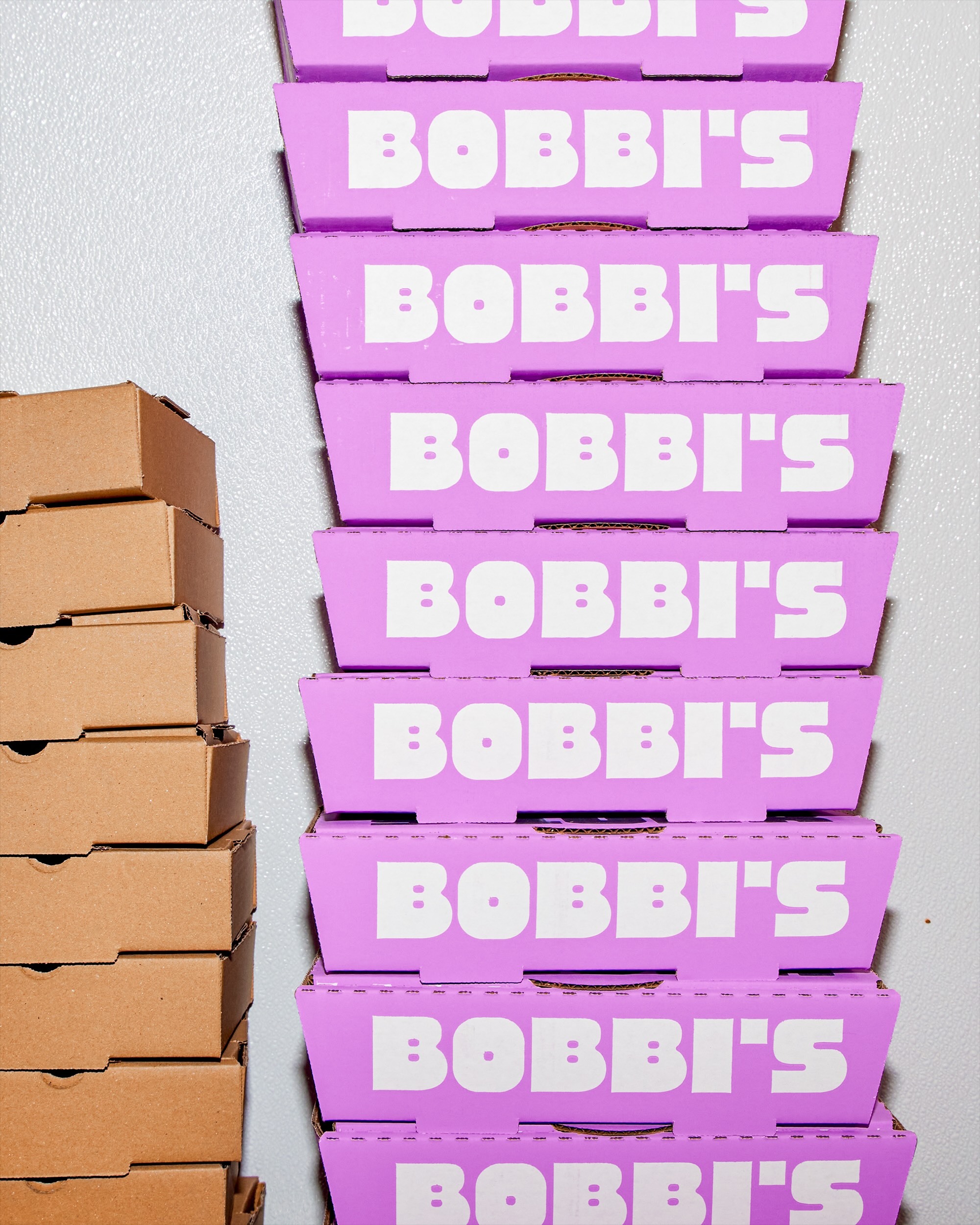 A towering stack of vibrant purple boxes with Bobbi's branding next to a stack of plain brown cardboard boxes against a light wall.