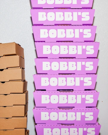 A towering stack of vibrant purple boxes with Bobbi's branding next to a stack of plain brown cardboard boxes against a light wall.