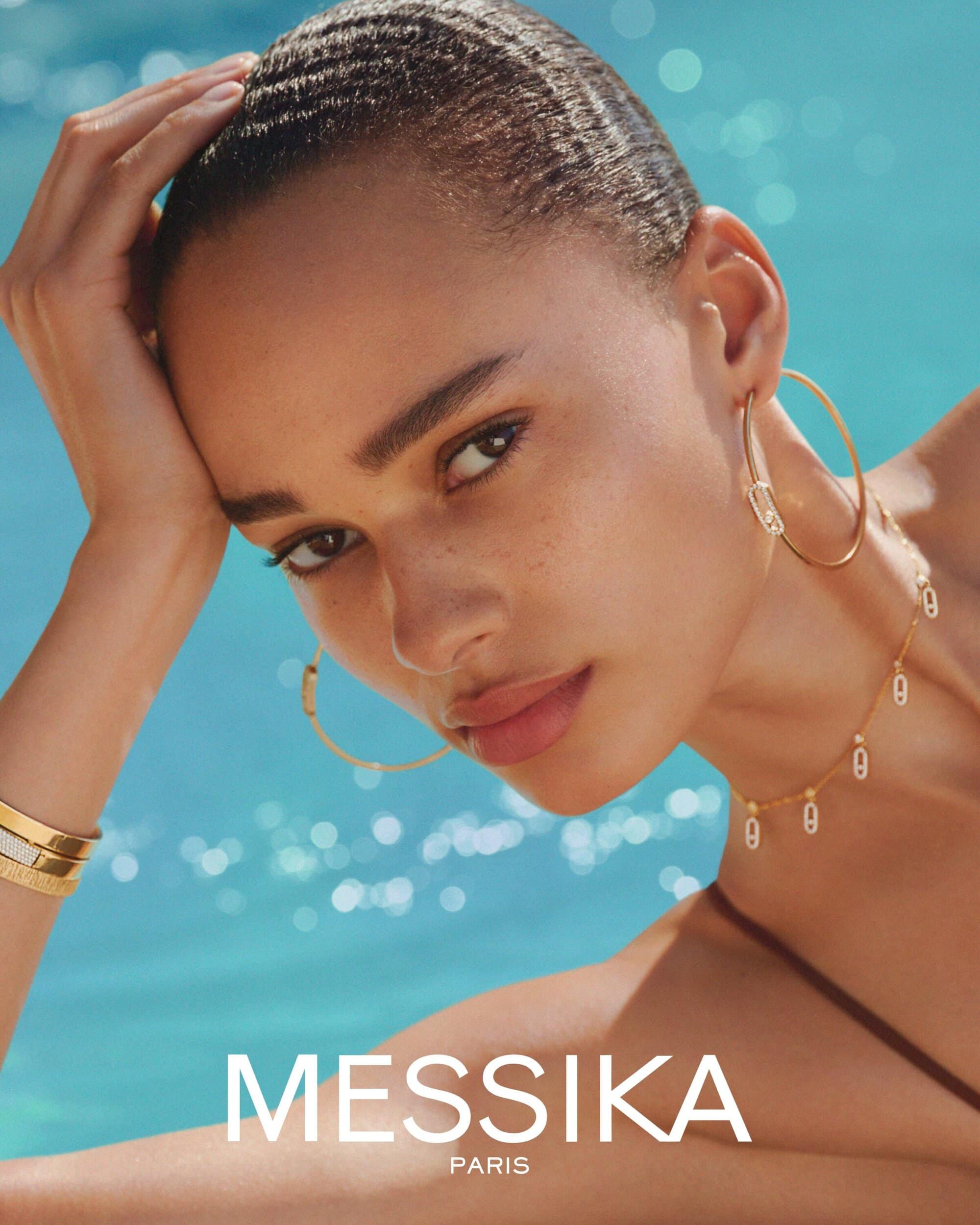 Close-up portrait of a model wearing gold hoop earrings and a delicate necklace against a bright blue pool water background