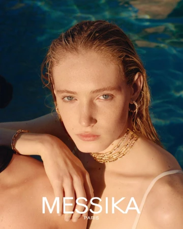 Close-up portrait of a woman with wet hair wearing gold chain necklaces and hoop earrings against a sparkling pool water background.