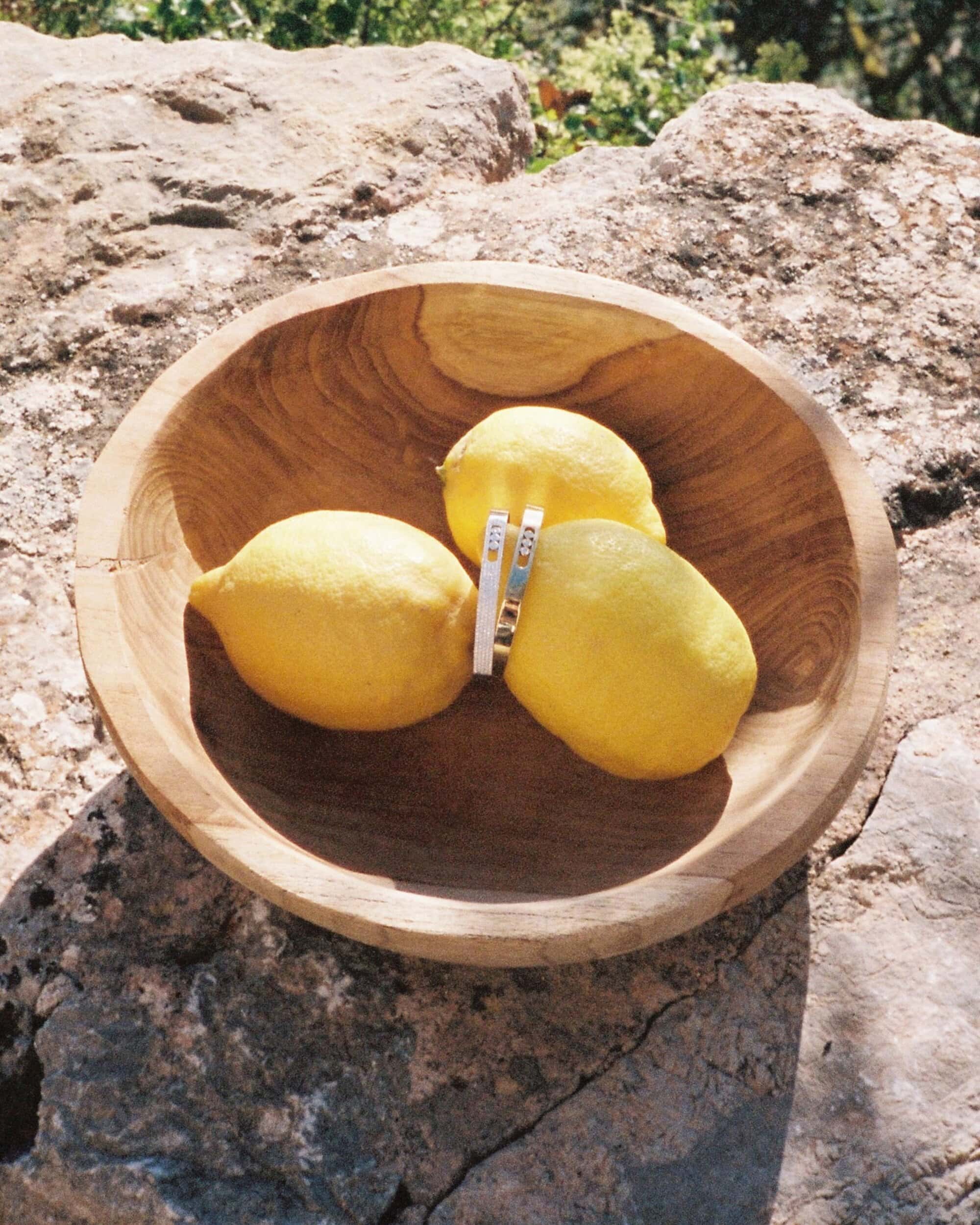 Three bright yellow lemons in a wooden bowl resting on a rough stone surface under bright sunlight.