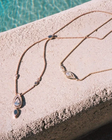 Close-up of delicate gold diamond necklaces resting on a textured stone ledge beside water for Messika.