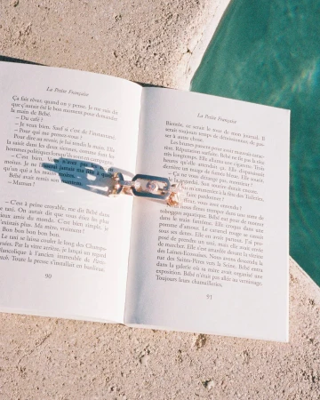 An open French-language paperback book resting on a stone surface beside sparkling turquoise water, with a gold ring placed on the pages.