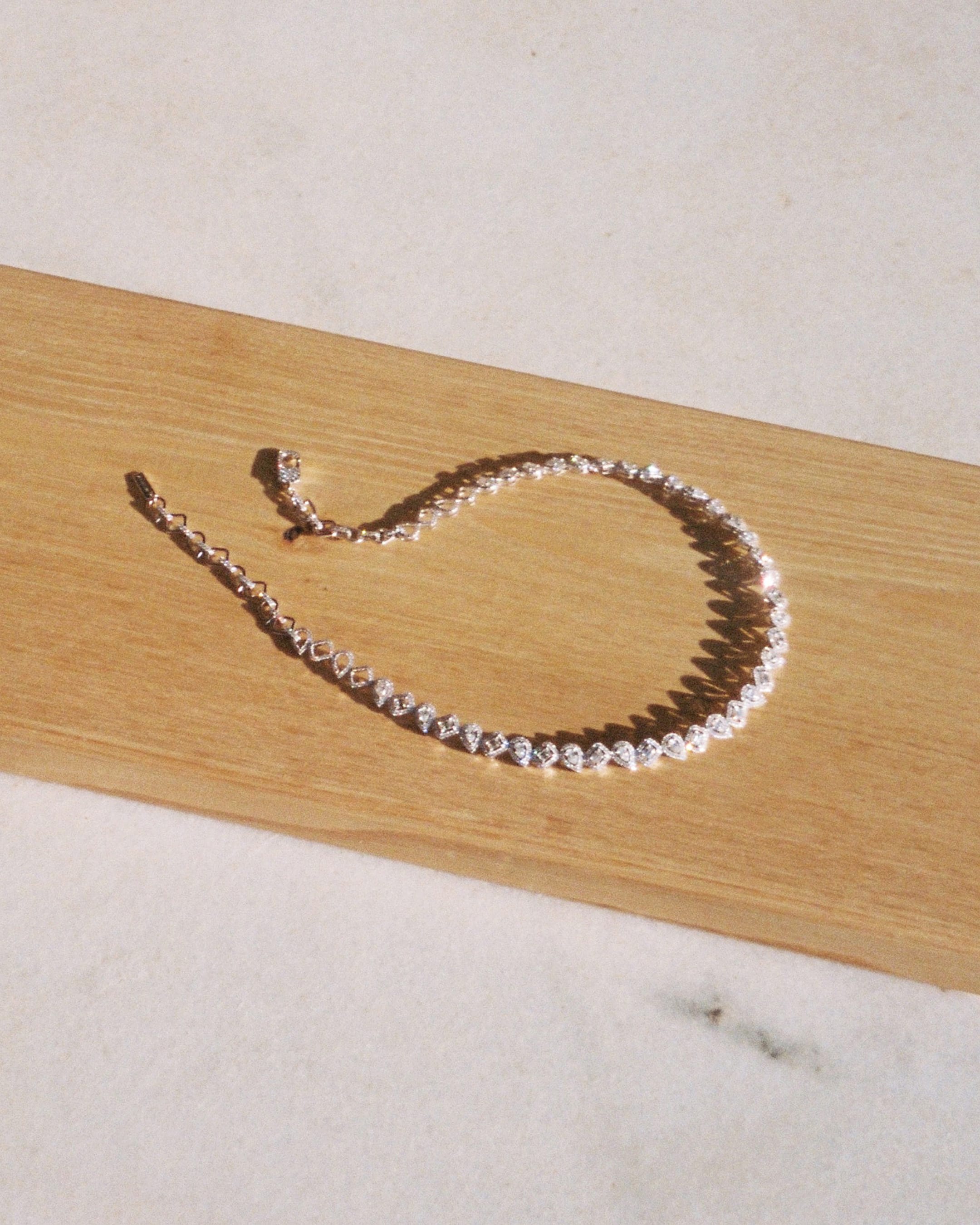 Sparkling diamond necklace resting diagonally on a textured wooden board and pale marble surface under direct sunlight.