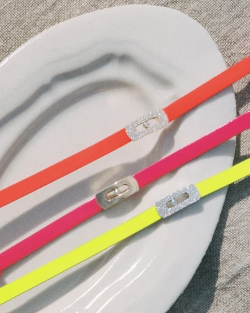 Three colorful neon bracelets with diamond clasps resting diagonally on a white ceramic dish against a textured linen background.