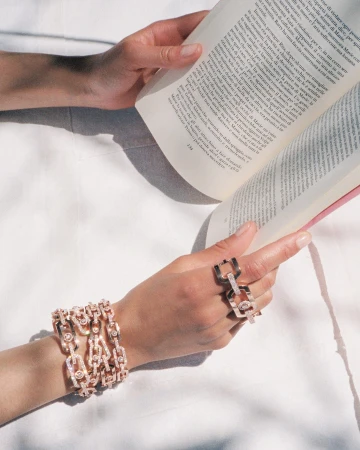 Close-up of hands holding an open book wearing diamond link bracelets and matching rings for Messika campaign.
