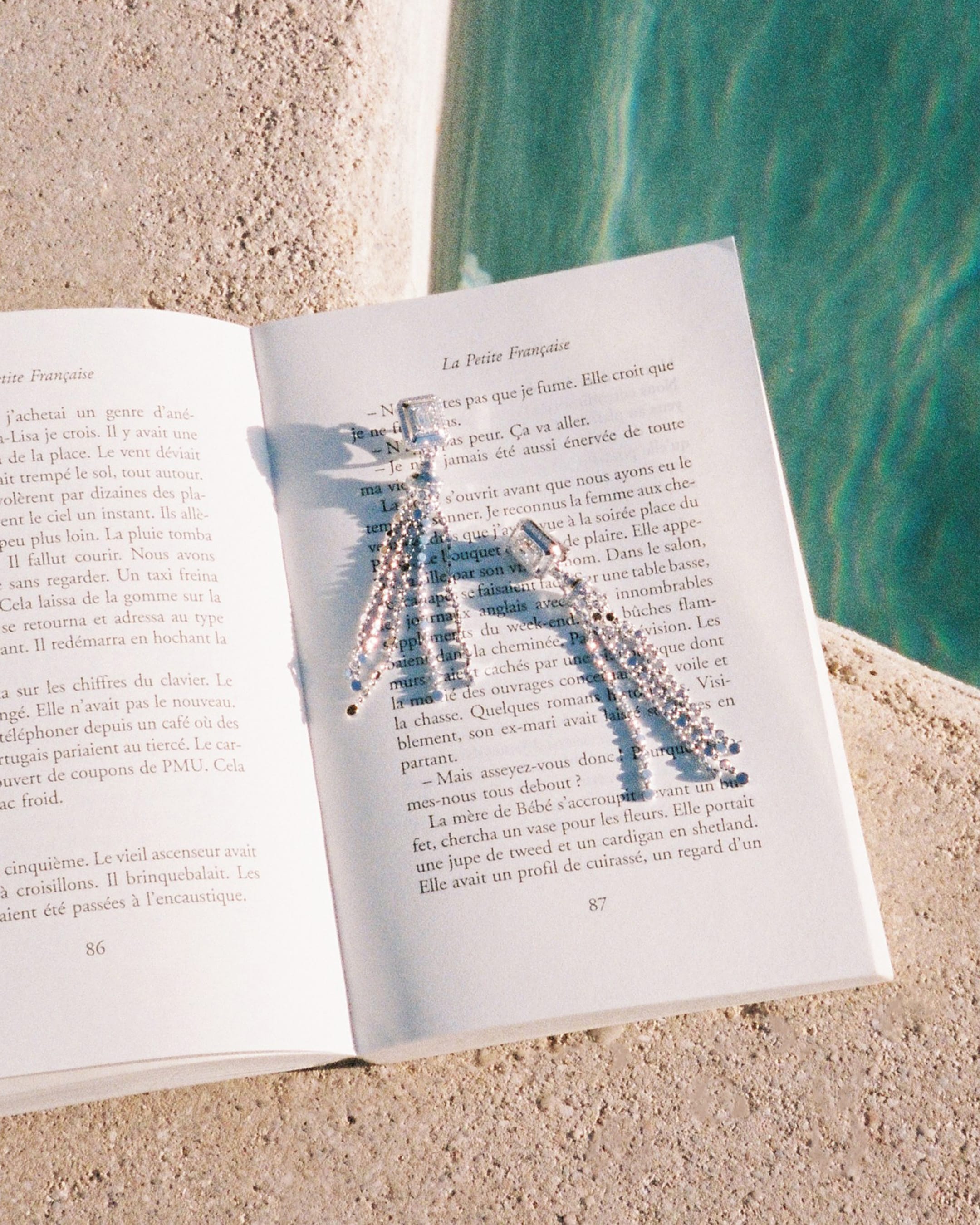 Diamond earrings resting on an open French book placed on a stone ledge beside a swimming pool.