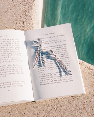 Diamond earrings resting on an open French book placed on a stone ledge beside a swimming pool.