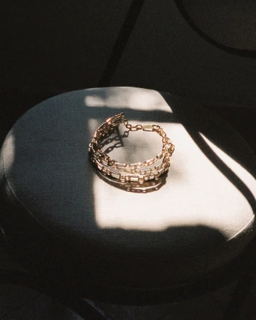 Gold chain bracelet resting on a round upholstered stool illuminated by harsh sunlight and shadows.