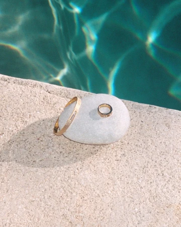 Gold jewelry including a ring and a bangle resting on a smooth white stone near turquoise swimming pool water