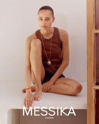 Model seated on a white ledge wearing a brown tank top and gold pendant necklace, photographed for Messika Paris.