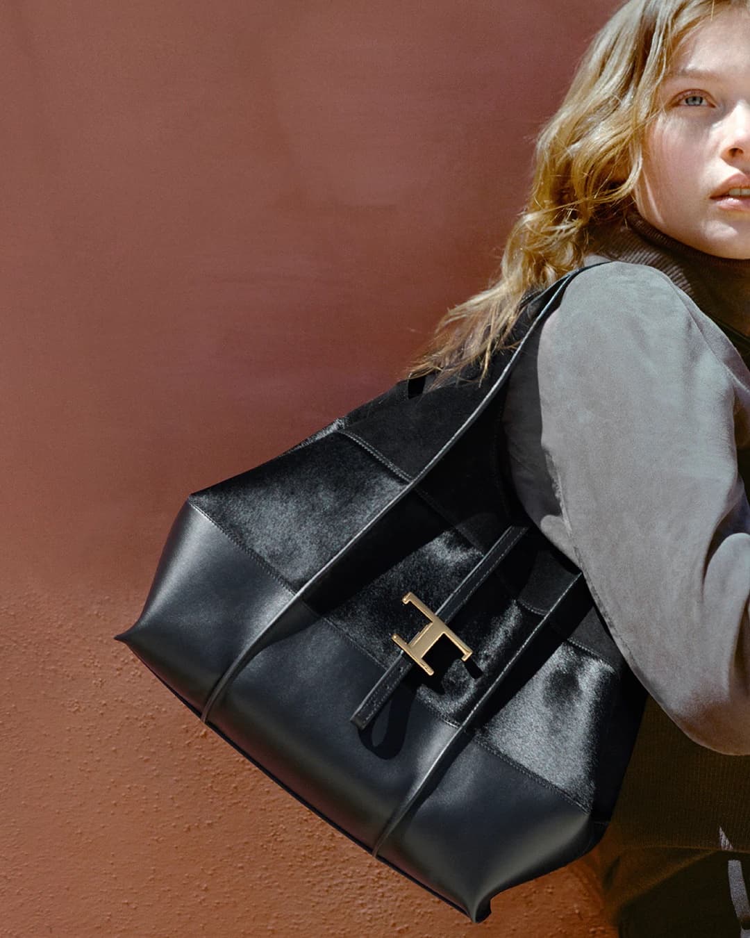 Close-up of a young woman carrying a large black leather shoulder bag against a warm terracotta wall.