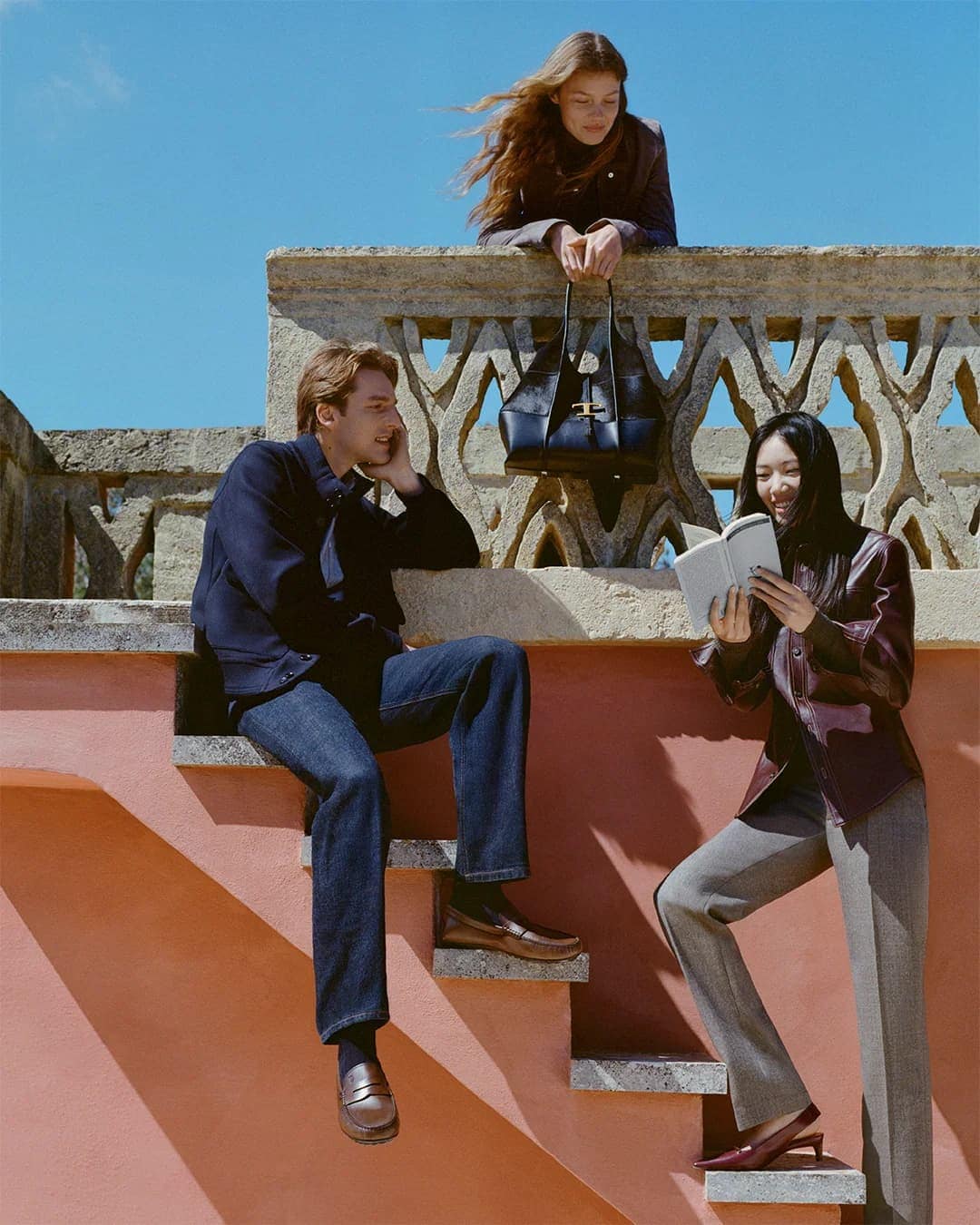 Three models posing outdoors on a stone staircase against a clear blue sky for Tod's Fall 2025 campaign.