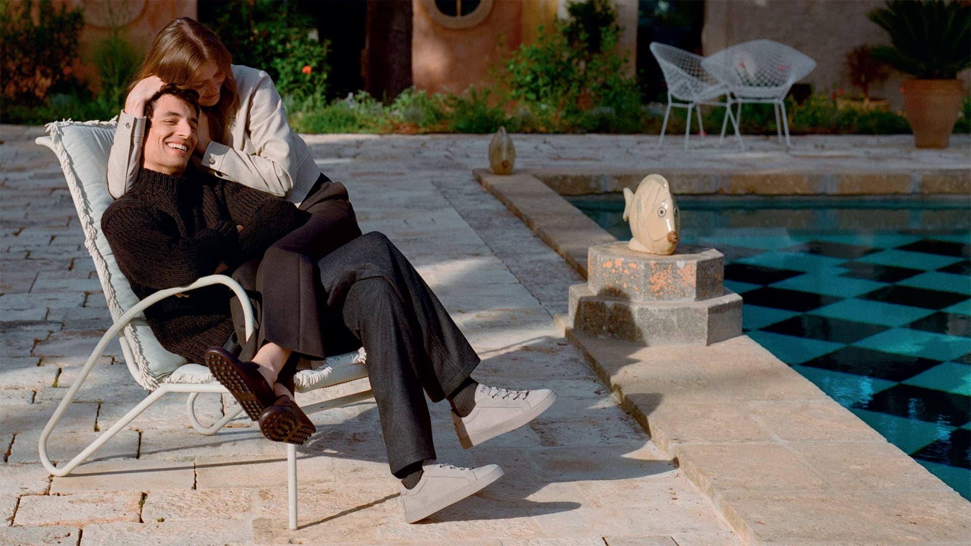 Man and woman sitting by a swimming pool, woman resting her hand on the man's head for Tod's Fall 2025 ad campaign.