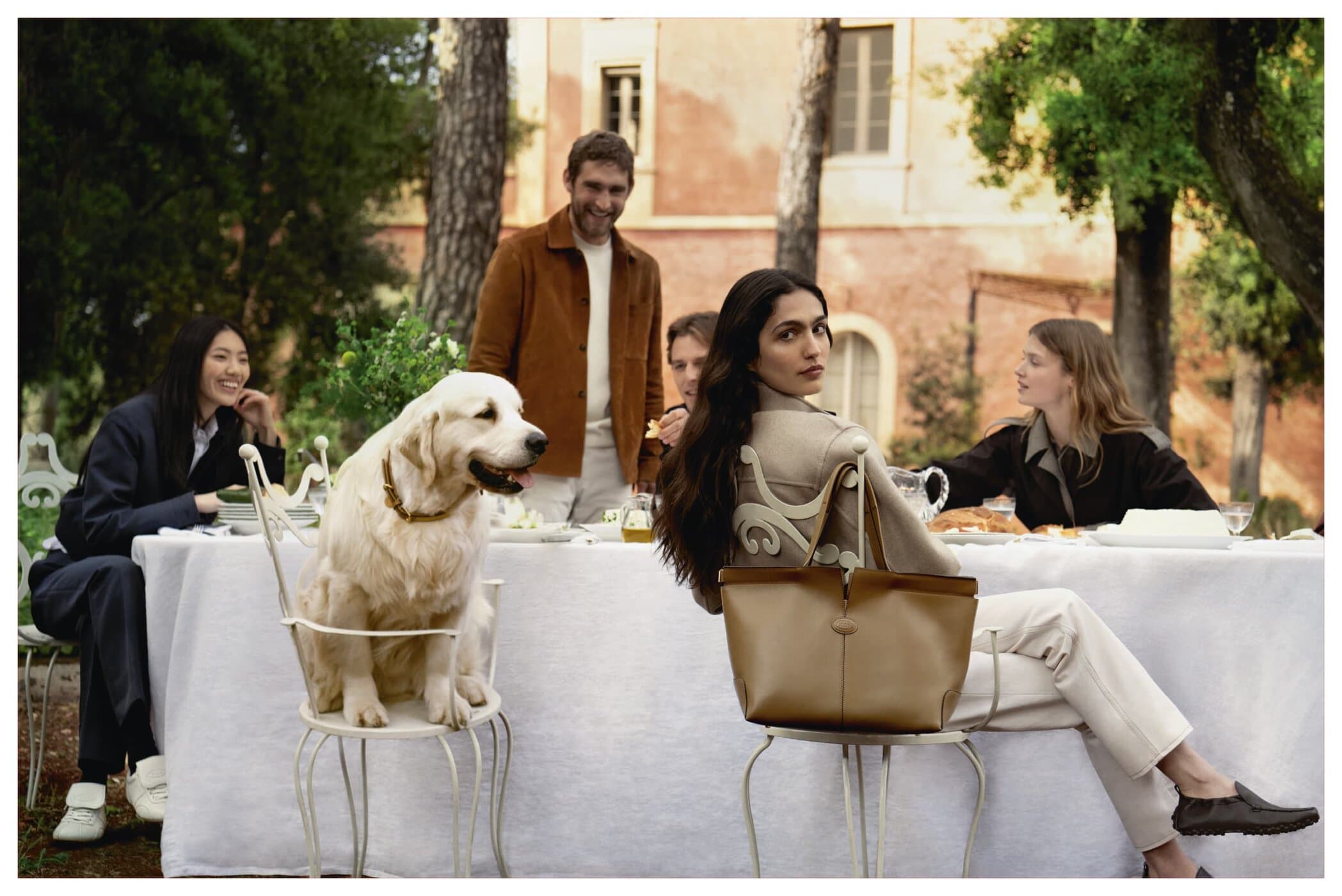 Group of people seated around a table outdoors with a golden retriever dog seated on a chair for Tod's ad campaign.