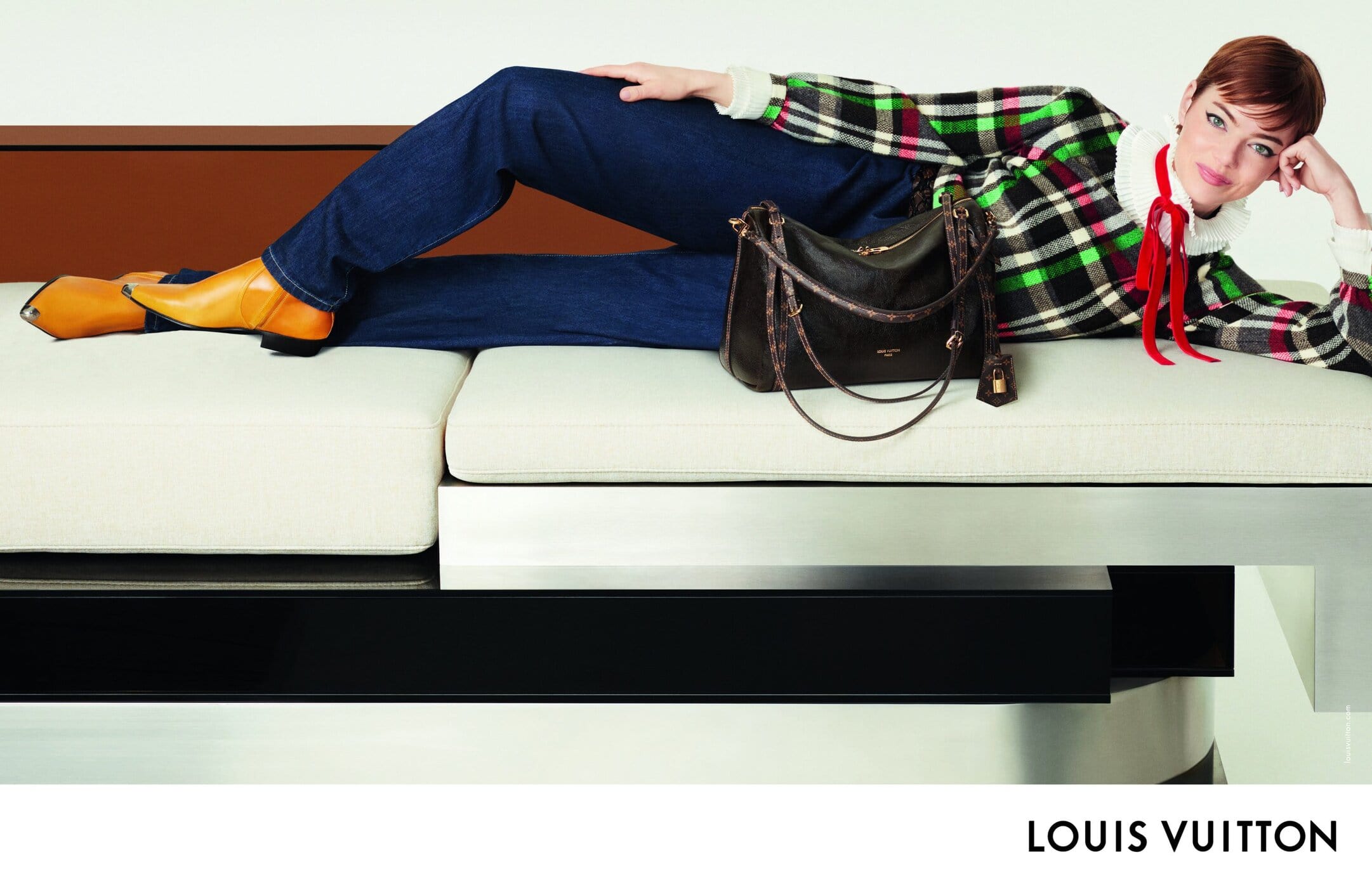 Emma Stone reposing on a modular white and metal bench wearing a plaid sweater, dark denim jeans, and yellow boots with a leather handbag