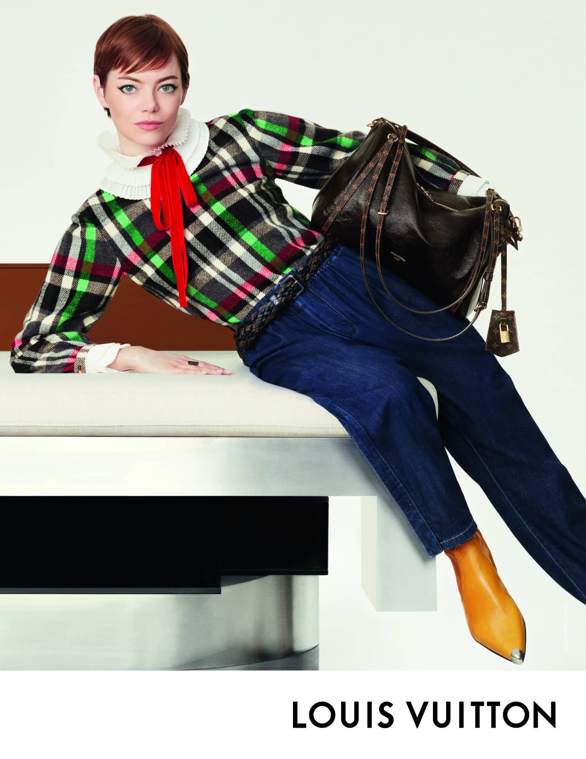 Emma Stone wearing plaid top and blue jeans leaning on furniture holding a dark shoulder bag for Louis Vuitton ad campaign