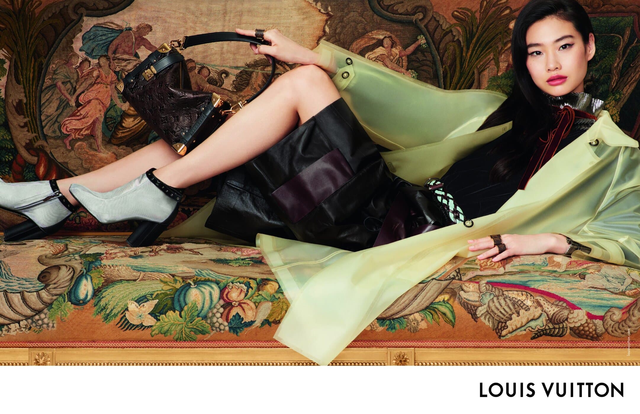 Model Hoyeon reclines on an ornate antique couch wearing a yellow-green translucent coat, holding a black Louis Vuitton handbag