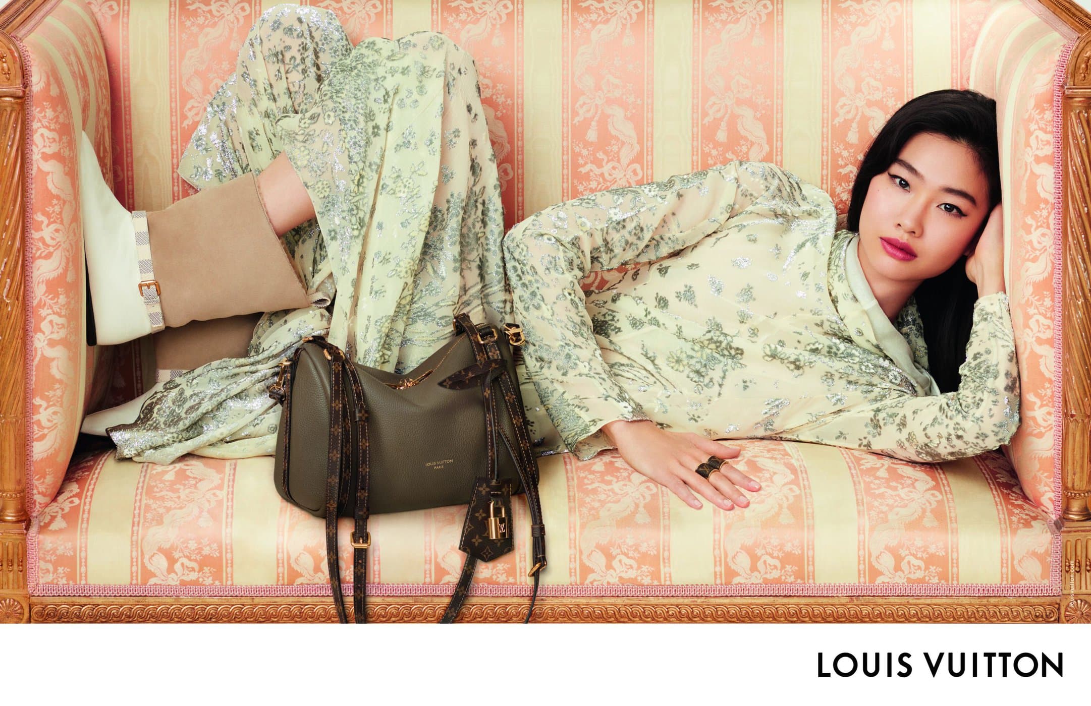 Model lying on an ornate pink and white striped sofa wearing a light green patterned dress alongside a dark green handbag.