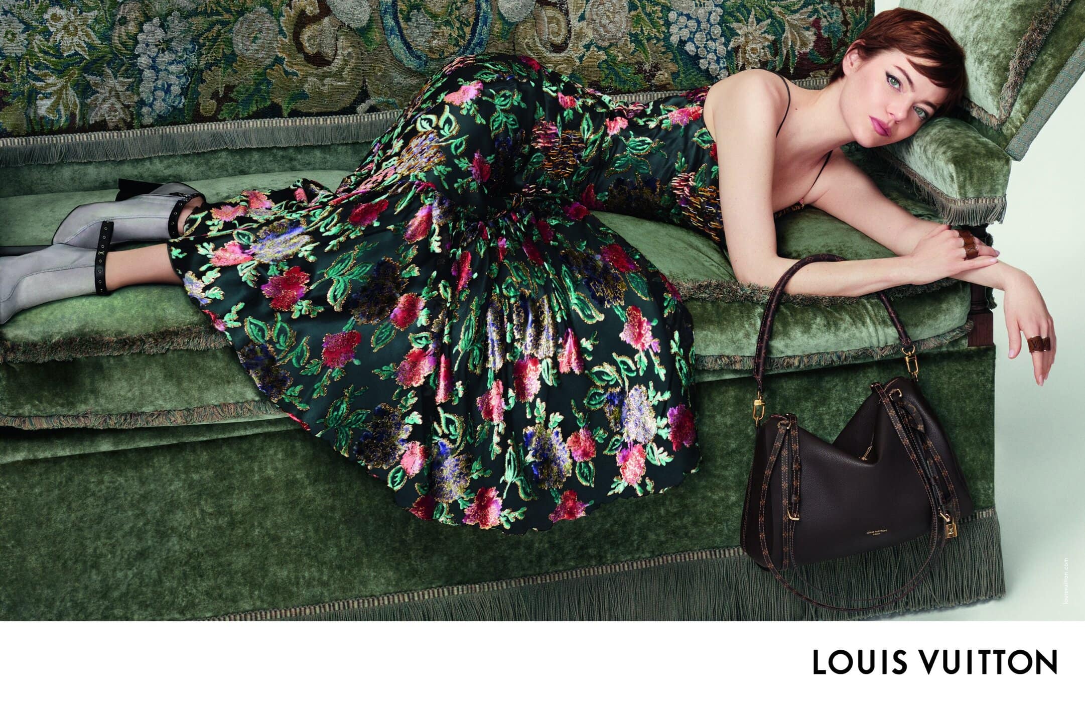 Emma Stone reclining on a green velvet sofa wearing a floral sequin dress with a dark leather handbag on the floor.