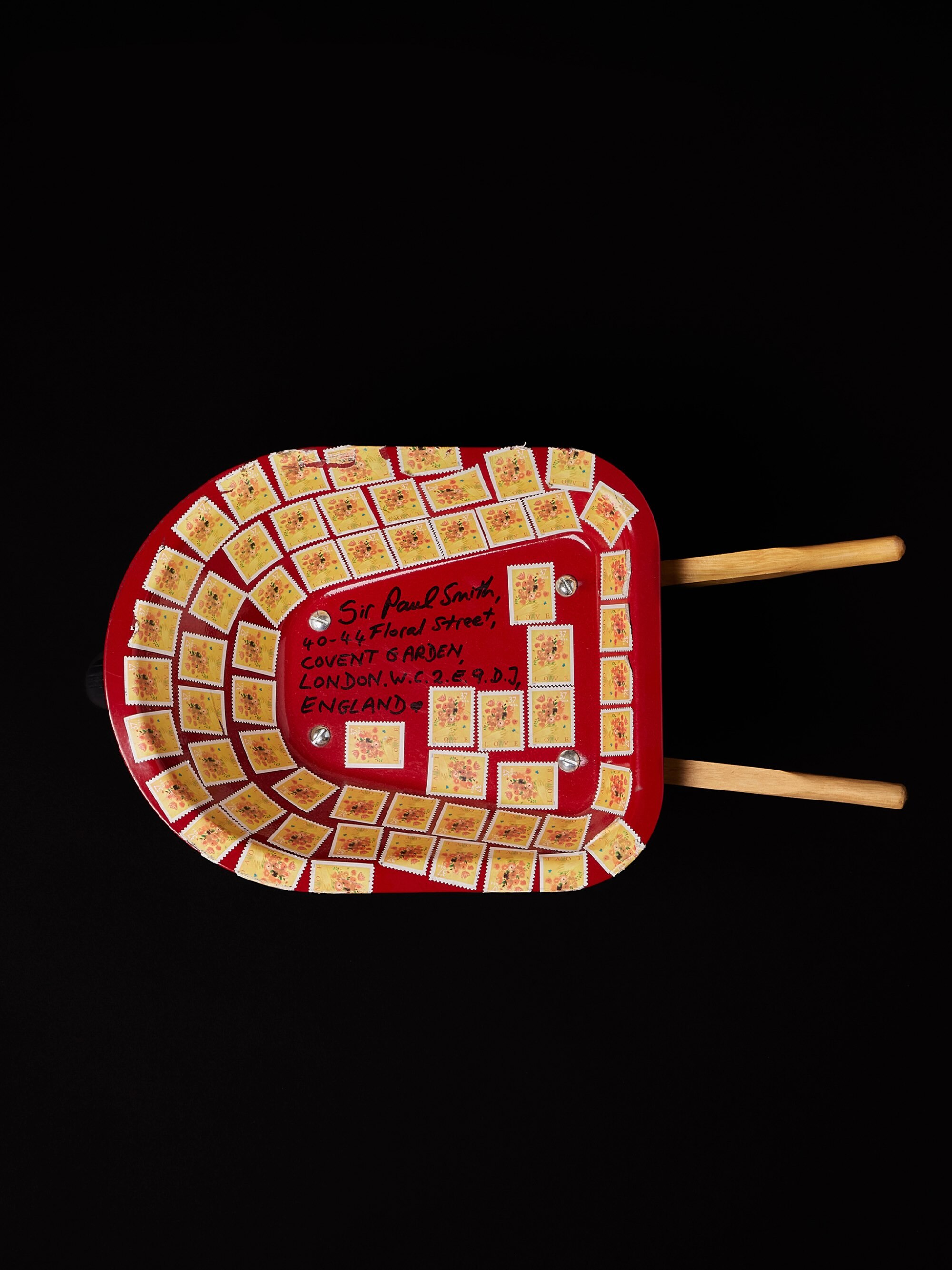 Red wheelbarrow decorated with postage stamps and handwritten address against a solid black background.