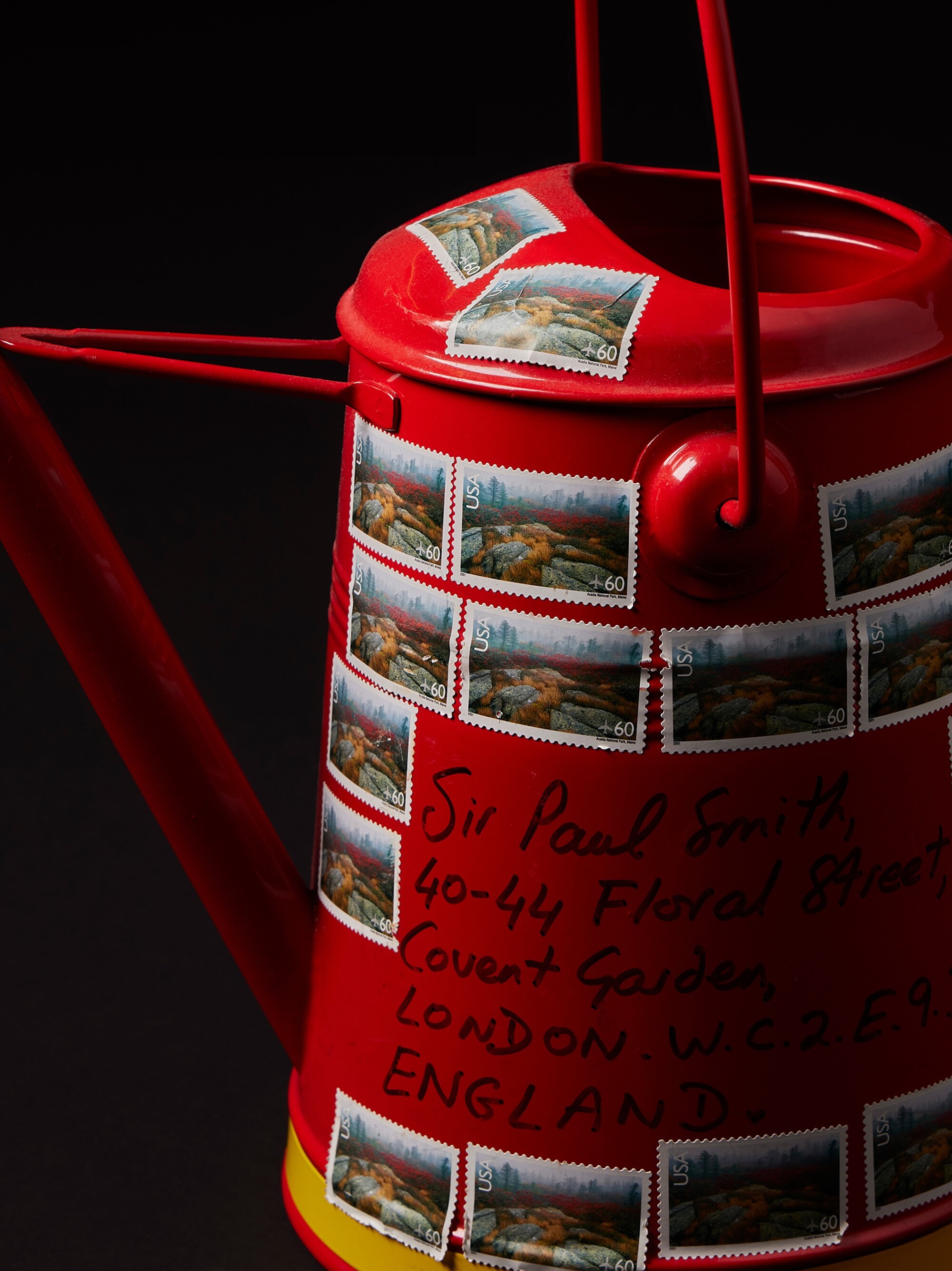 Bright red metal watering can covered in postage stamps and handwritten address text against a dark background