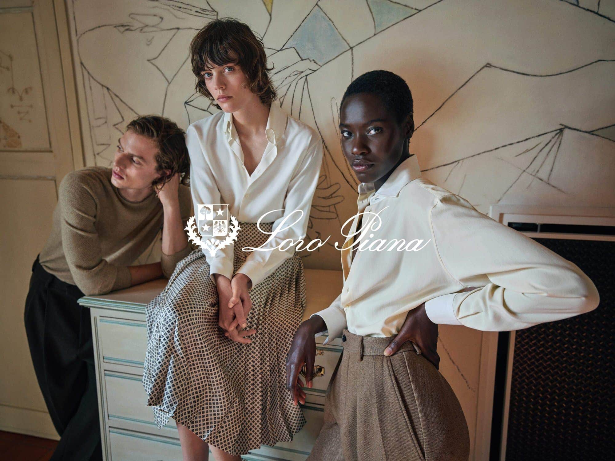 Three models posing indoors before an illustrated wall panel for the Loro Piana Fall 2025 campaign shot by Mario Sorrenti.
