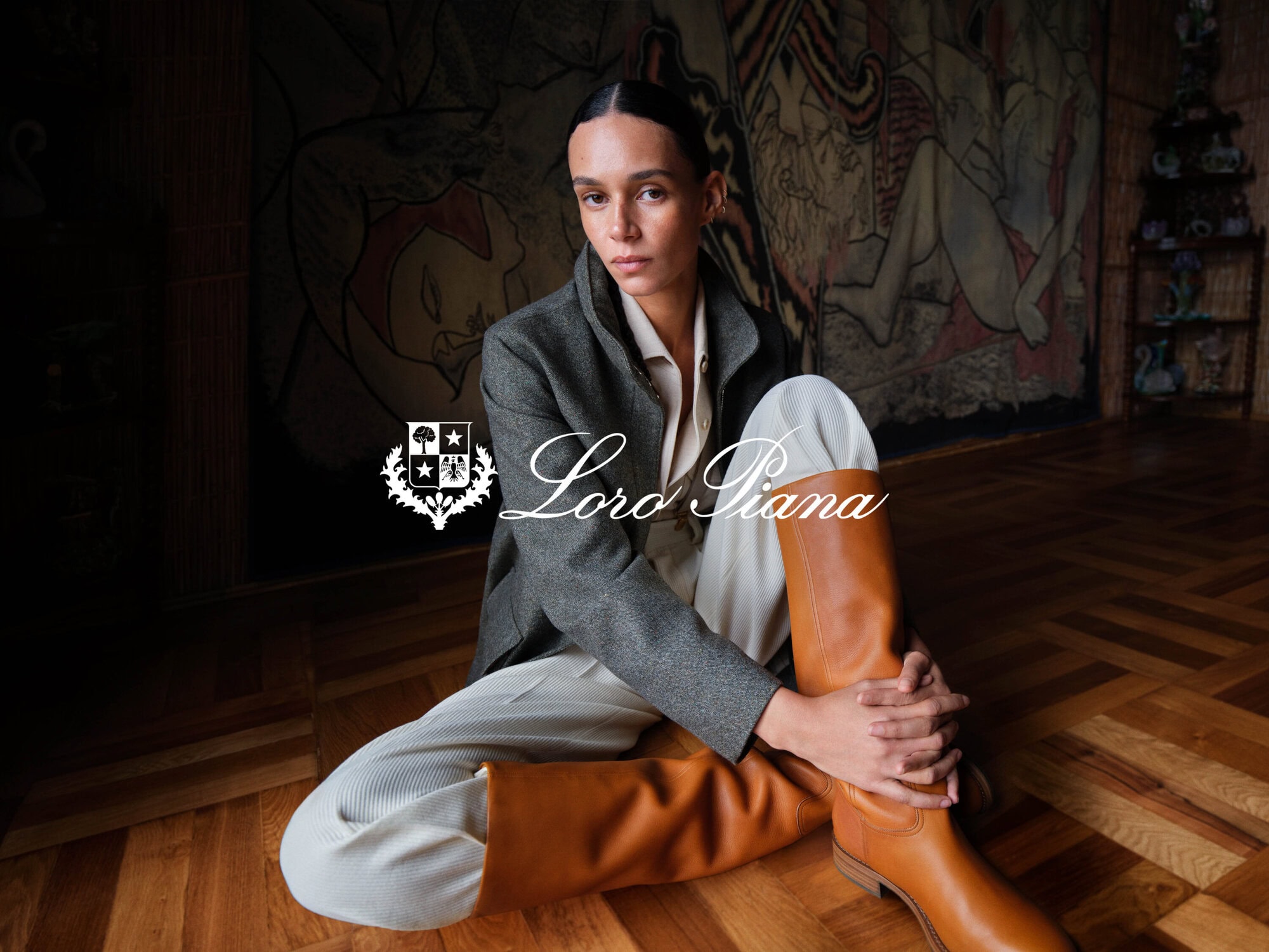 Model seated on a wooden floor wearing a grey jacket, light trousers, and tall tan leather boots with a wall mural background