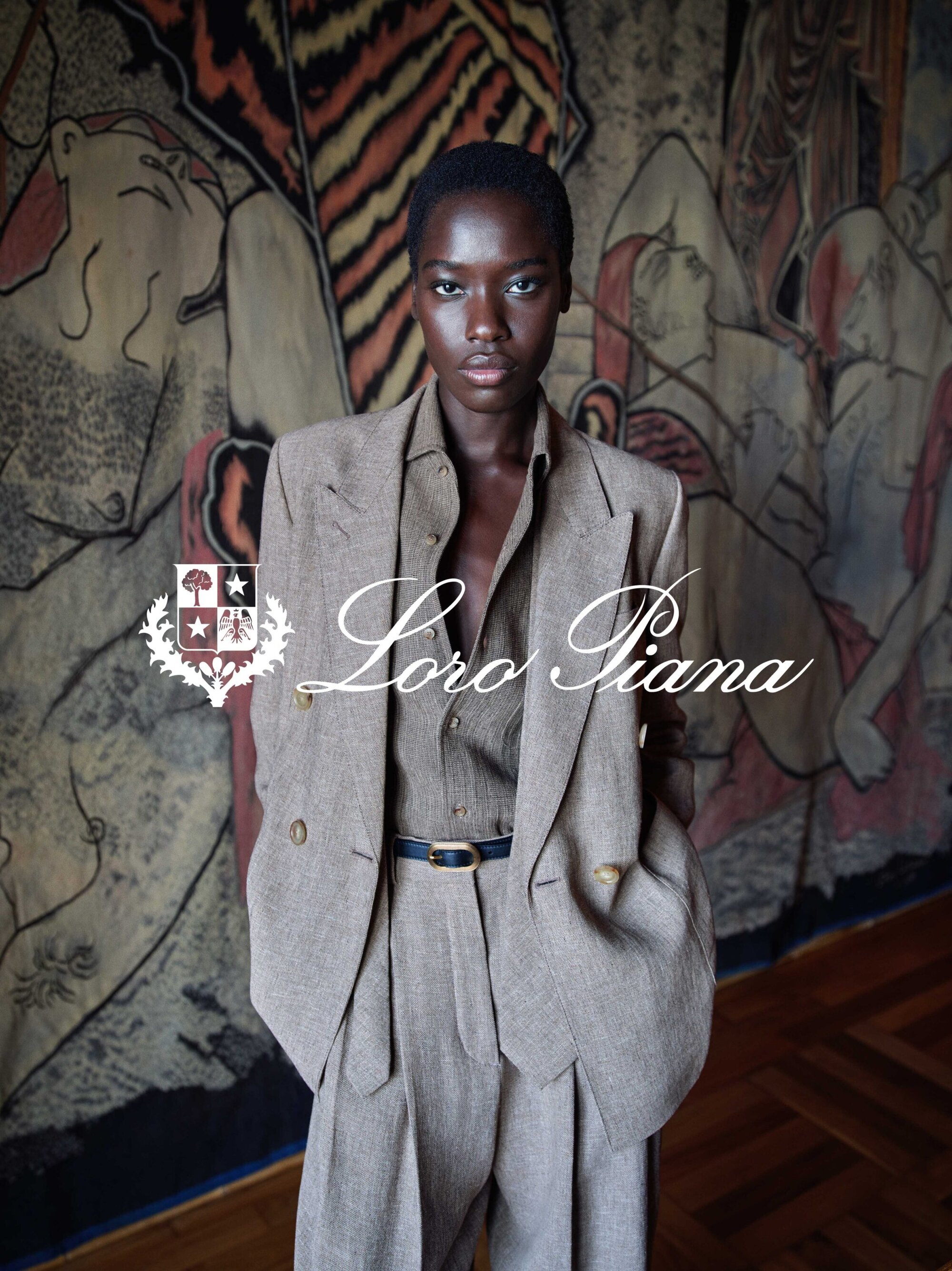 A model wearing a tailored beige suit and shirt stands before a large illustrated wall tapestry for a Loro Piana ad campaign.