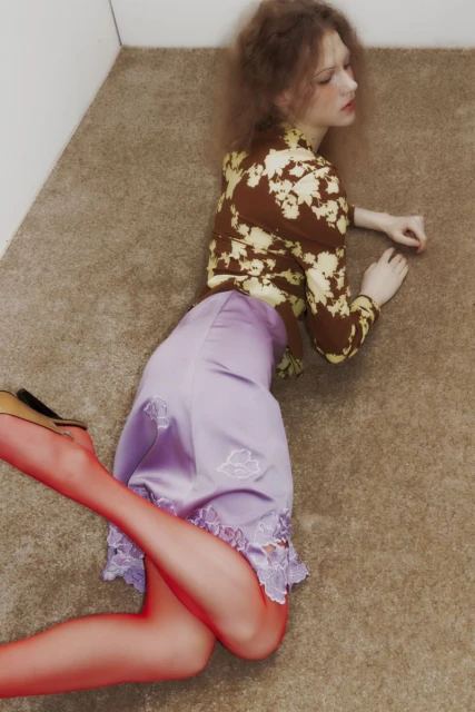 A woman lies on a carpeted floor wearing a patterned blouse, lilac skirt with lace trim, sheer orange hosiery and tan-colored heels.