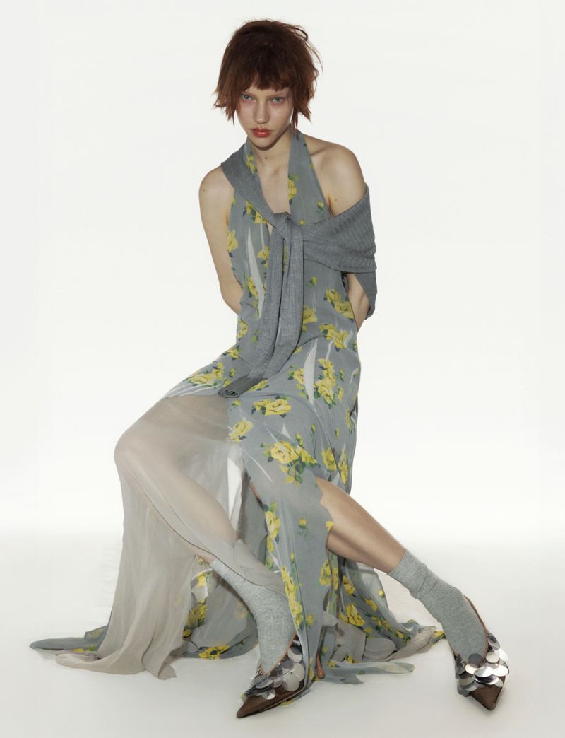 A woman with short auburn hair poses in a light gray floral dress with metallic, pointed-toe shoes against a white backdrop.