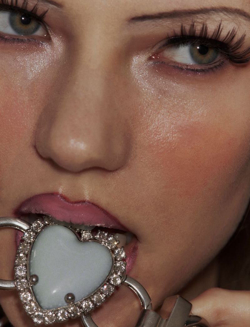 Close-up of a young woman with dramatic eye makeup biting a large, decorative heart-shaped object near her pink-glossed lips.