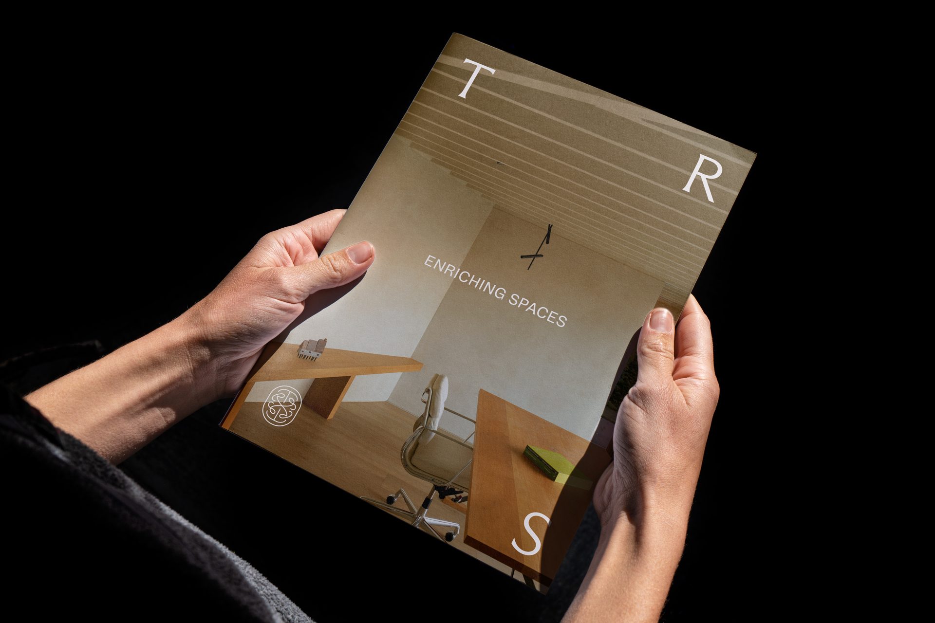 Two hands holding a printed brochure titled Enriching Spaces for TR Studio against a dark background
