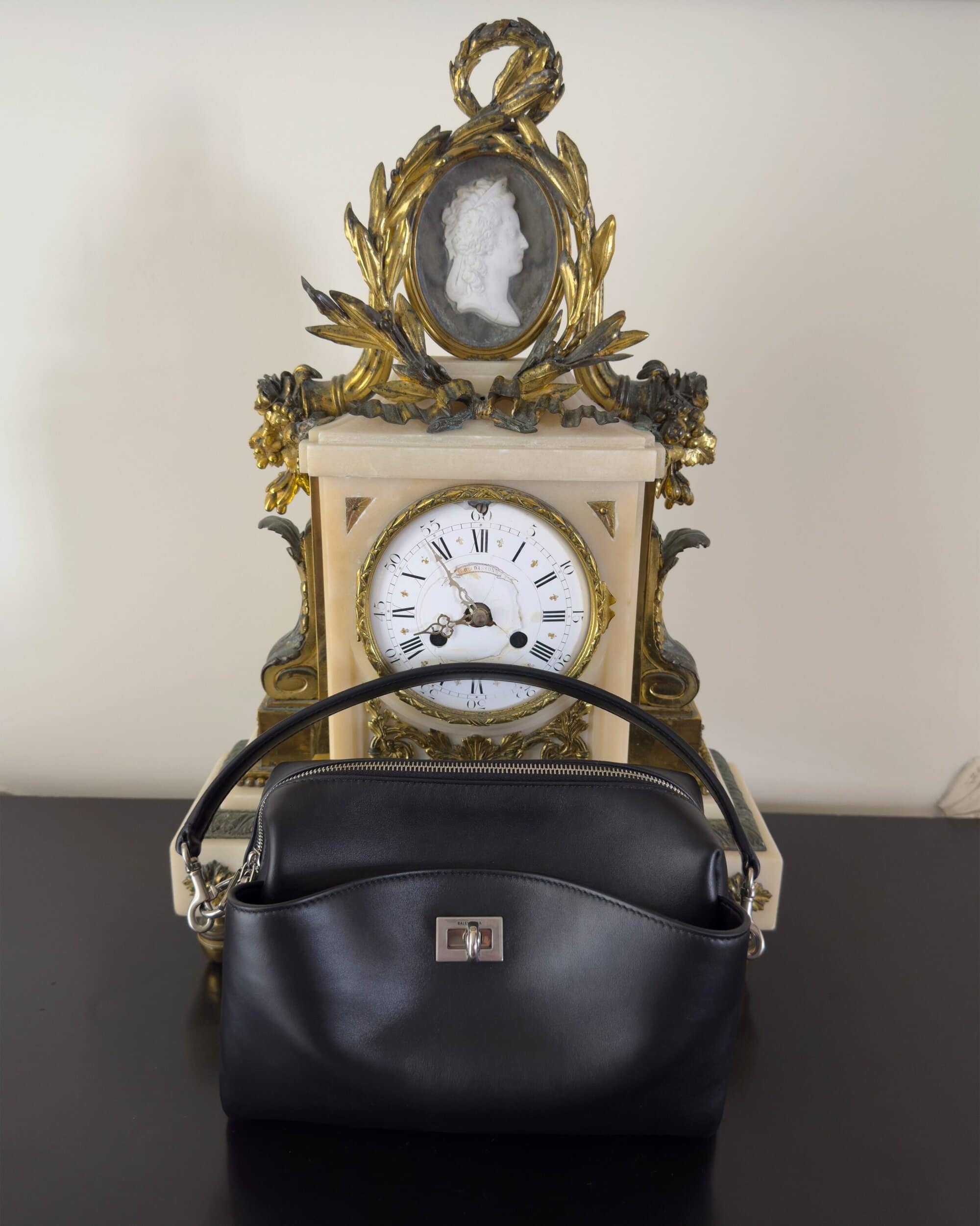 Black leather handbag positioned on a dark surface in front of an ornate antique clock with a cameo portrait