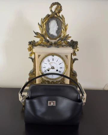 Black leather handbag positioned on a dark surface in front of an ornate antique clock with a cameo portrait