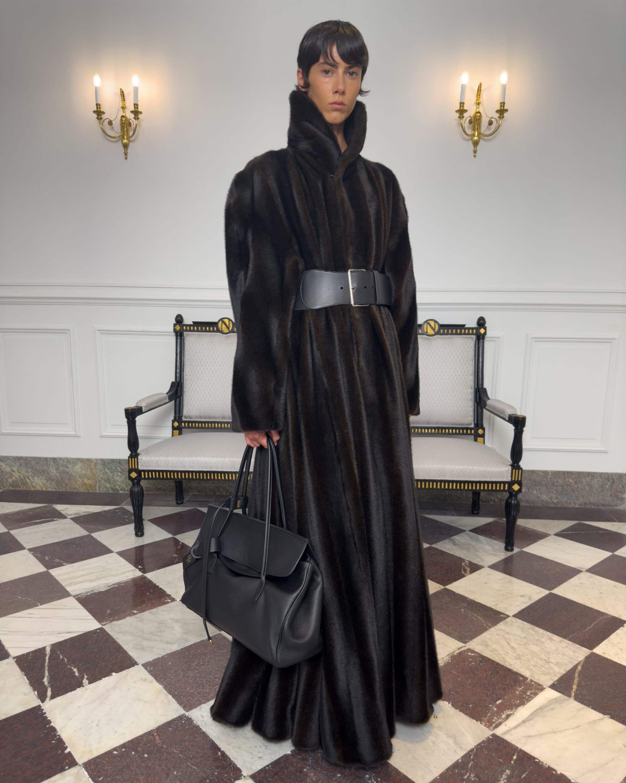 Model standing on a checkered marble floor wearing a long dark fur coat and holding a black leather handbag