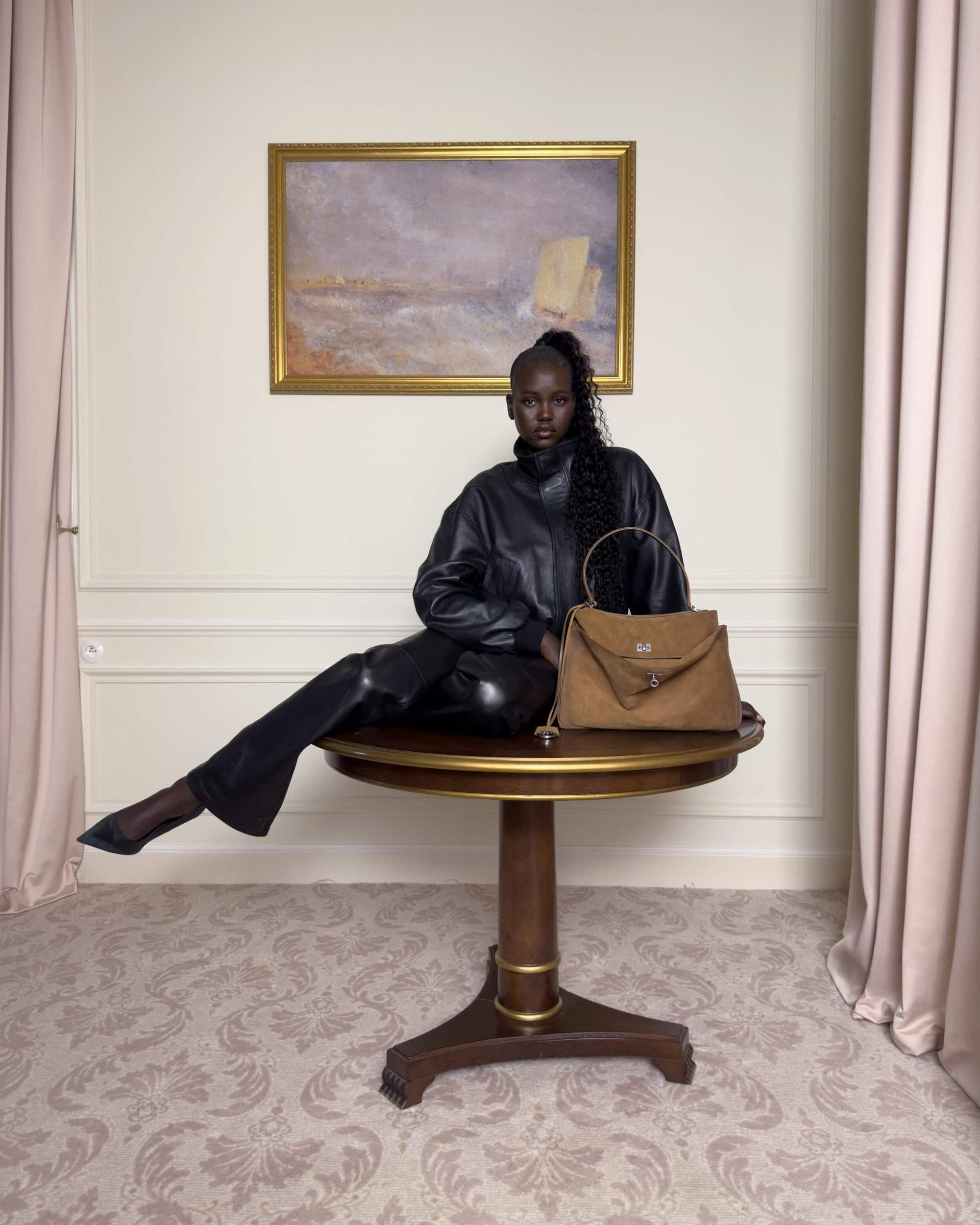 Model wearing black leather jacket and pants posed on a wooden round table holding a tan handbag