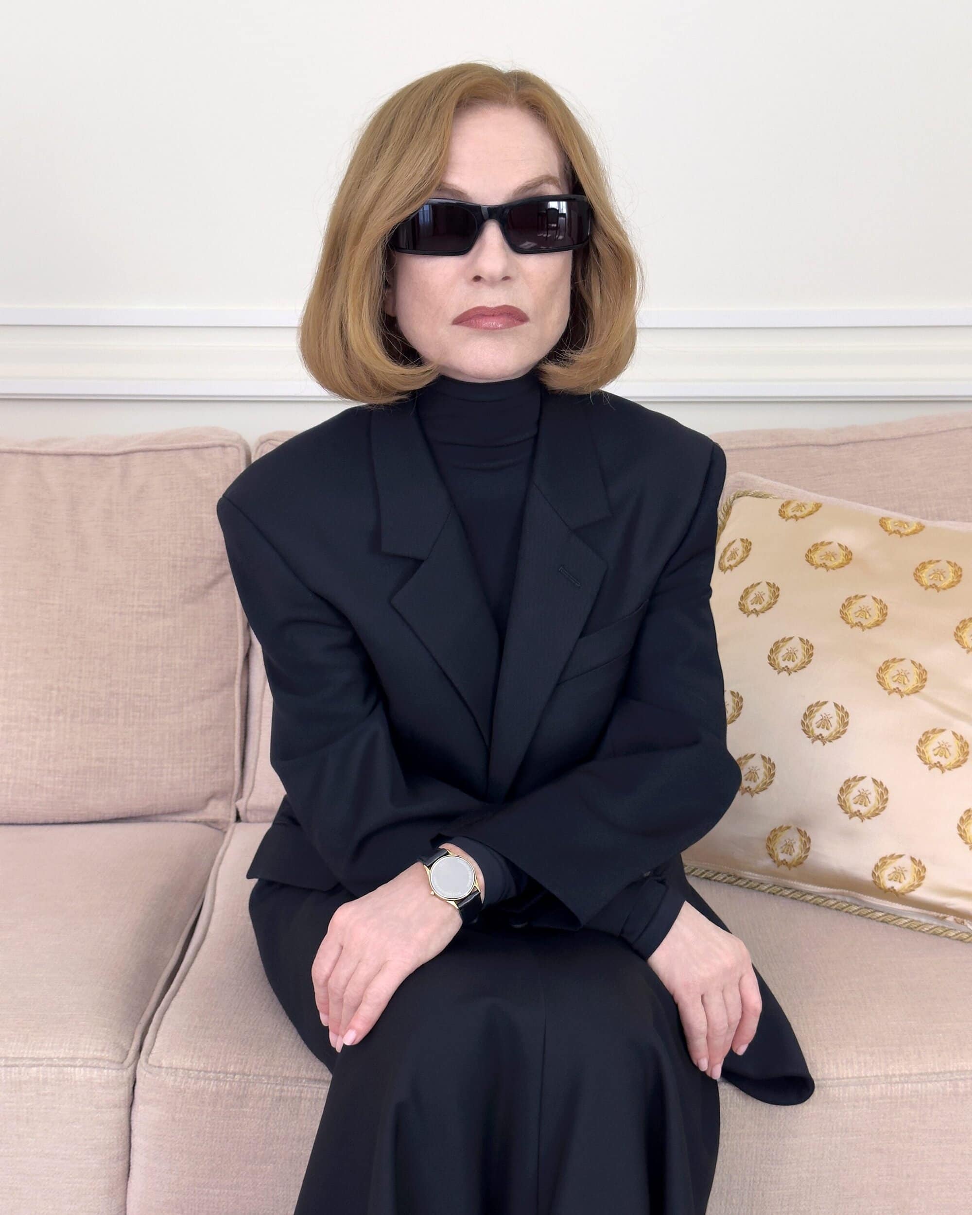 Isabelle Huppert wearing black sunglasses and a dark suit seated on a pink sofa for Balenciaga