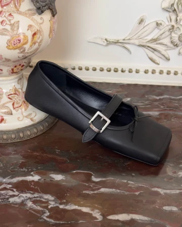 Black Mary Jane shoe with silver buckle resting on a dark marble surface beside a decorative ceramic vase.