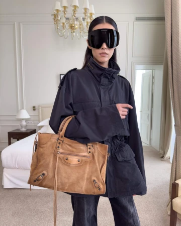 Model wearing dark oversized jacket and wrap-around sunglasses holding a large brown leather handbag indoors