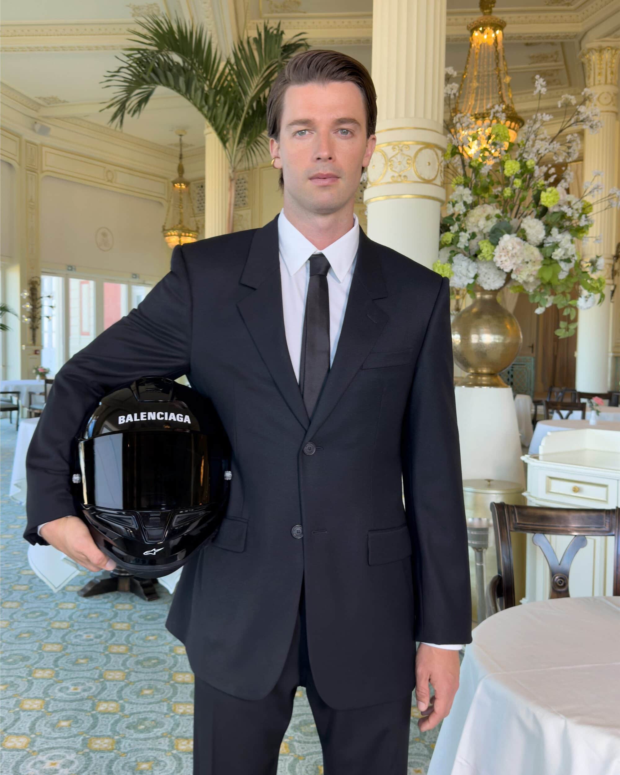 Man wearing a black suit and white shirt holding a branded black motorcycle helmet inside a hotel interior